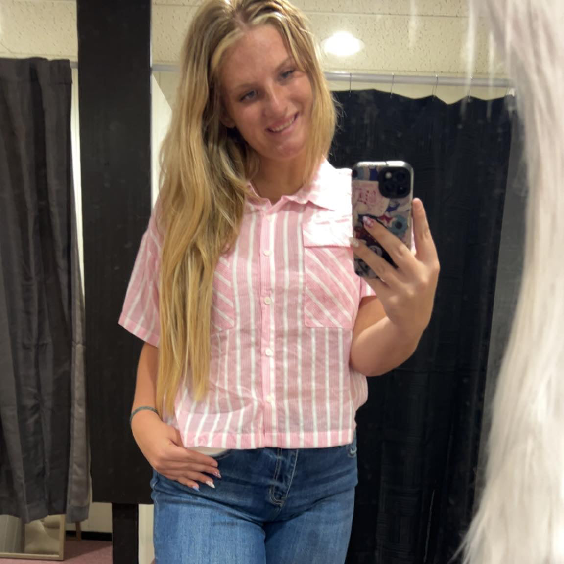Person taking a mirror selfie wearing a pink striped shirt and blue jeans.